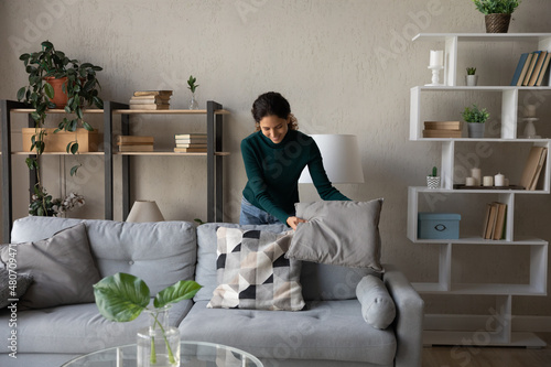 Happy millennial generation positive beautiful young housewife cleaning apartment, arranging stuff in cozy modern living room, put things on right places, feeling satisfied with comfort tidy house. photo