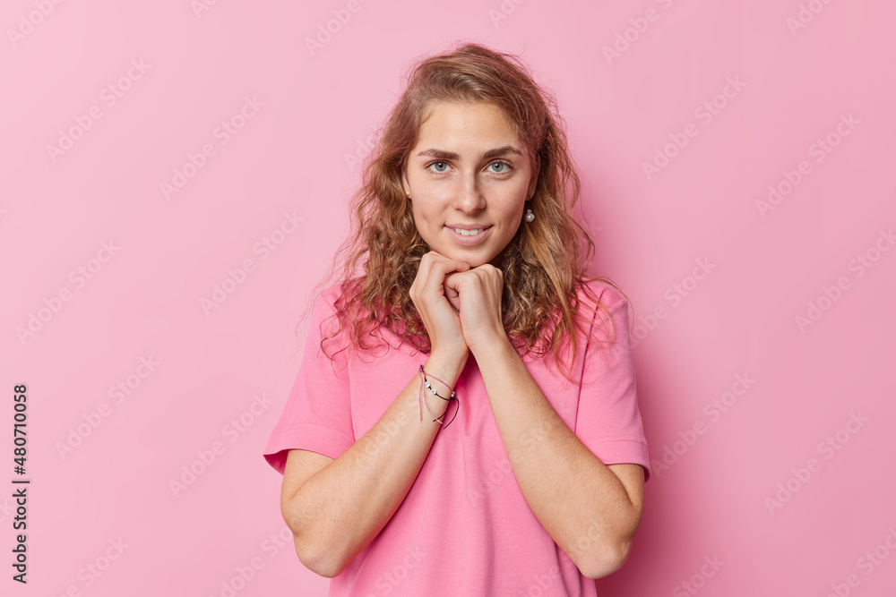 Charming beautiful blue eyed young woman with long wavy hair keeps hhands under chin looks directly at camera dressed in casual basic t shirt isolated over pink background poses for making photo Charming beautiful blue eyed young woman with long wavy hair keeps hhands under chin looks directly at camera dressed in casual basic t shirt isolated over pink background poses for making photo