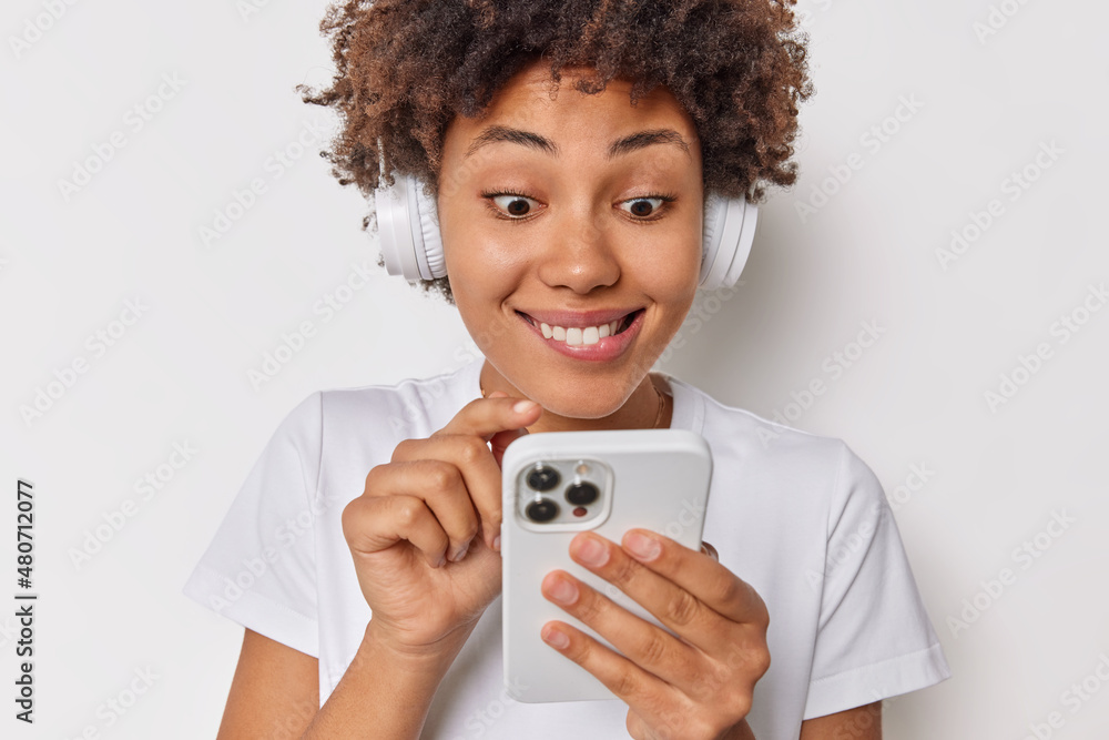 Happy curious curly haired woman bites lips looks at smartphone scrolls newsfeed listens music ia headphoes dressed casually isolated over white background. People and modern technologies concept Happy curious curly haired woman bites lips looks at smartphone scrolls newsfeed listens music ia headphoes dressed casually isolated over white background. People and modern technologies concept