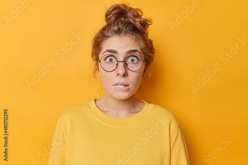 Fotografie Embarrassed nervous woman with anxious expression bites lips stars through round spectacles finds out something dressed in casual jumper isolated over yellow background
