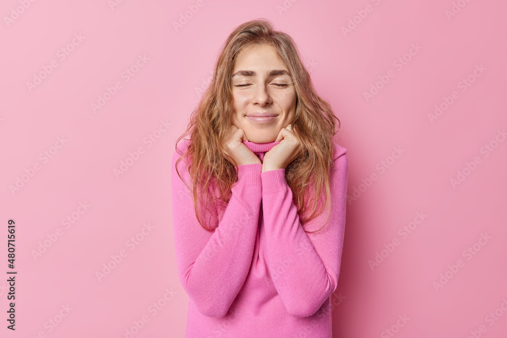 Tender beautiful young woman keeps hands under chin closes eyes dreams about something pleasant has long hair enjoys softness of jumper isolated over pink background. People and daydreaming. Tender beautiful young woman keeps hands under chin closes eyes dreams about something pleasant has long hair enjoys softness of jumper isolated over pink background. People and daydreaming.