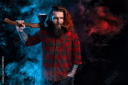 Bearded lumberjack. Brutal man with cigar in checkered shirt with long hair holds old axe in smoke