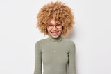 Portrait of good looking young woman with curly bushy hair smiles toothily gazes directly at camera wears spectacles and turtleneck feels positive isolated over white background. Emotions concept