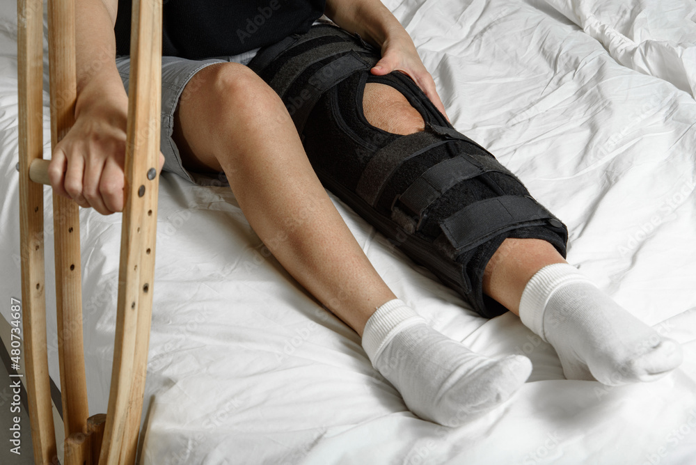 Female patient wearing orthosis sitting on bed holding crutches after surgery on broken leg. Rehabilitation of injured woman at home. Selective focus.