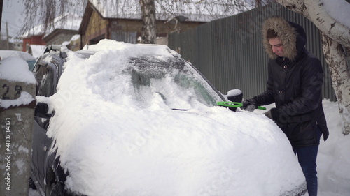 A young man in a jacket cleans his car of snow with a brush. The car was covered in snow. Weather disaster.