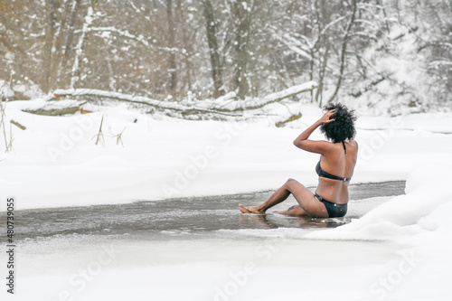 Beautiful girl sitting on the ice the cold water of a lake or river, cold therapy, ice swim with winter landscape and forest on background