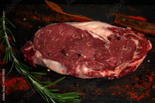 Raw ribeye steak with seasonings and a knife. A piece of marbled beef with rosemary and a mixture of peppercorns. Black background. Space for text.