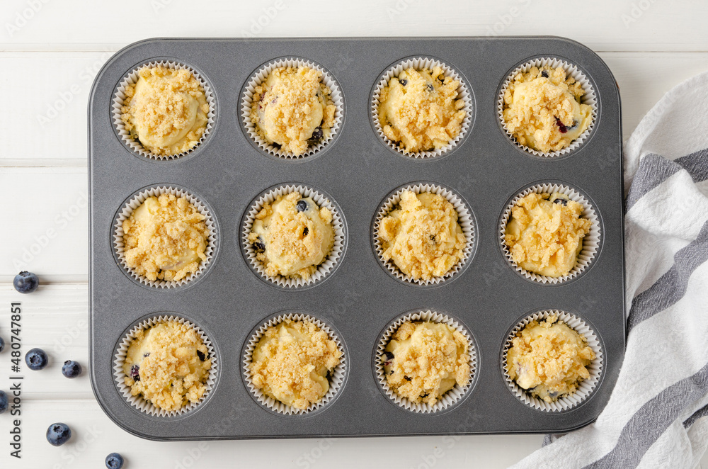 Fototapeta premium Raw dough for lemon muffins with blueberries and shtreisel in baking form on a white wooden background. Recipe step by step. Top view.