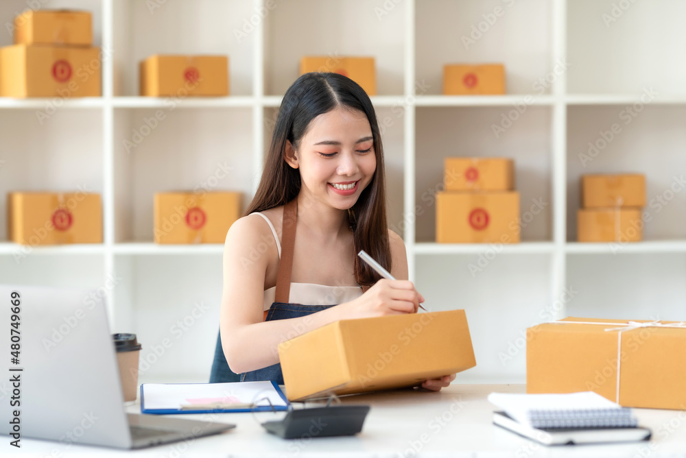 Online sales young Asian business owner woman is taking note of the customer's address on the parcel box being prepared for delivery.