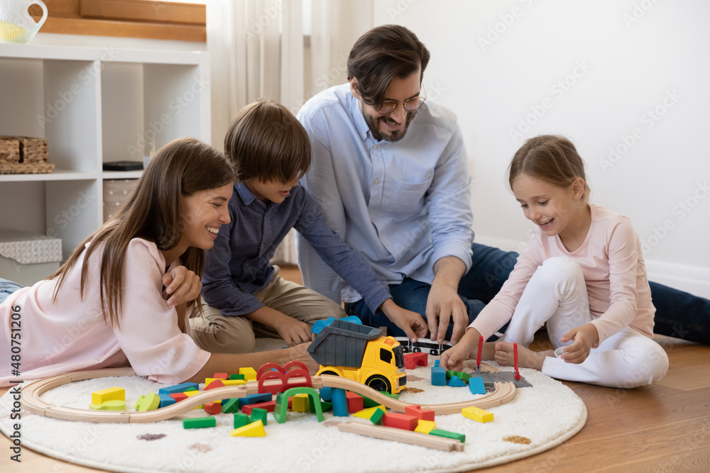 Couple and children spend time together sit on warm floor with underfloor heat enjoy playtime playing wooden railroad and colorful bricks. Quarantine leisure of family with kids, toys store ad concept Couple and children spend time together sit on warm floor with underfloor heat enjoy playtime playing wooden railroad and colorful bricks. Quarantine leisure of family with kids, toys store ad concept