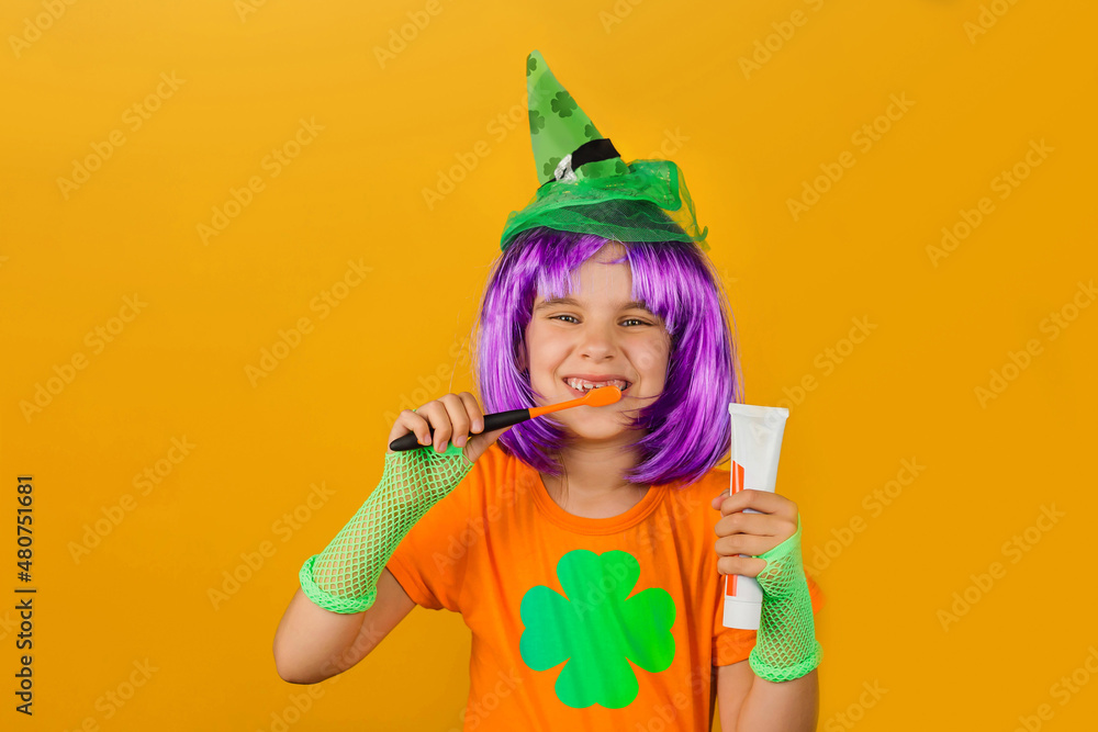 St.Patrick 's Day. Child girl a carnival costume leprechaun green wig, hat  holds with toothpaste and an orange brush isolated on a yellow background.  Medicine, dental hygiene, holidays concept. Stock Photo