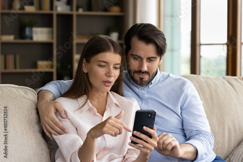 Beautiful millennial couple in love sit on sofa with smartphone, look at device screen discussing purchase, choose goods items for house using e-services at home. Modern tech, new mobile app concept