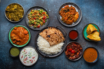Assorted Indian ethnic food buffet on rustic concrete table from above: curry, fried samosa, rice biryani, dal, paneer, chapatti, naan, chicken tikka masala, mango lassi, dishes of India for dinner