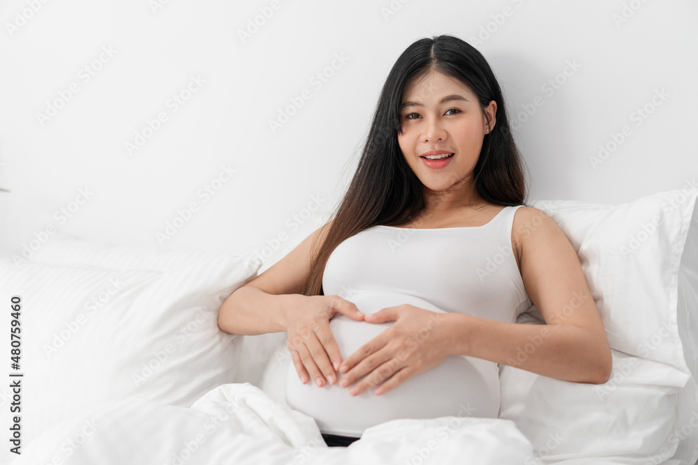 Asian happy pregnant woman is sitting on bed and and touching her belly. pregnancy, motherhood, people and expectation concept Asian happy pregnant woman is sitting on bed and and touching her belly. pregnancy, motherhood, people and expectation concept