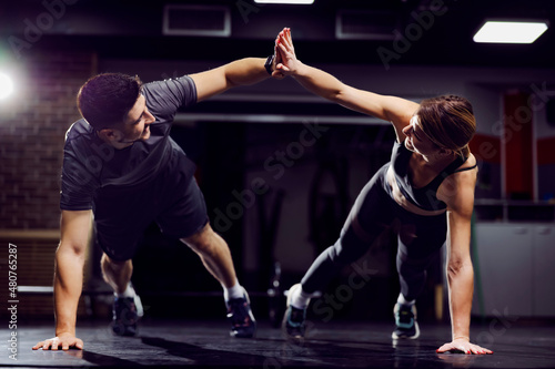 A fit sporty couple giving high five to each other while doing planks and exercises in a gym.