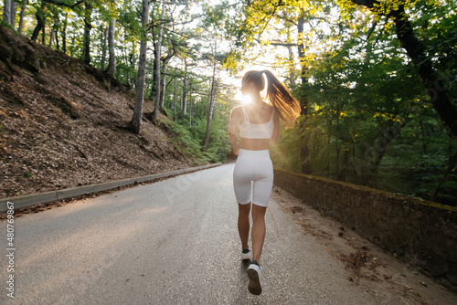 A young beautiful girl in white sports clothes is running with her back, on the road in a dense forest, during sunset. Doing sports in the fresh air. A healthy lifestyle.