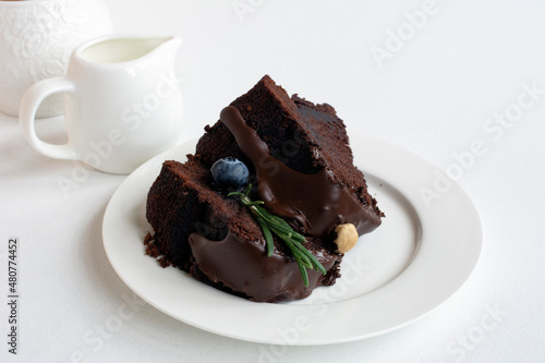 Chocolate cake with icing, decorated with fresh blueberry and green rosemary twig, on plate on table with sugar bowl and creamer  on light background