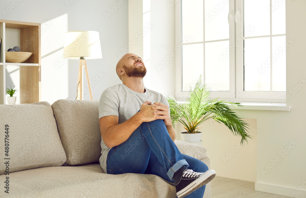 Millennial man with rheumatism, arthritis or osteoarthritis experiences severe pain in knee joint. Unhappy suffering man grimaces from terrible knee pain while sitting on sofa at home. Health concept. Millennial man with rheumatism, arthritis or osteoarthritis experiences severe pain in knee joint. Unhappy suffering man grimaces from terrible knee pain while sitting on sofa at home. Health concept.