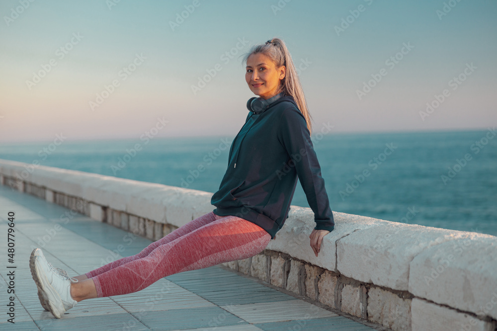 Side view of healthy woman in sportswear warming up before exercising on the seafront, smiling at camera