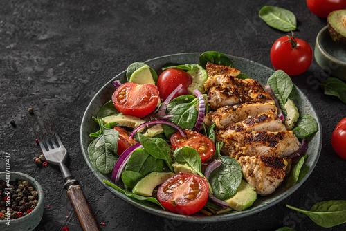 Grilled chicken breast fillet and spinach salad with avocado, tomatoes and sesame seeds in a plate on dark background