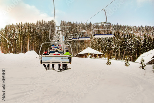 Snowboarder and skiers ride on a ski lift to a snowy mountain on a sunny day. Snowy forest with wooden houses