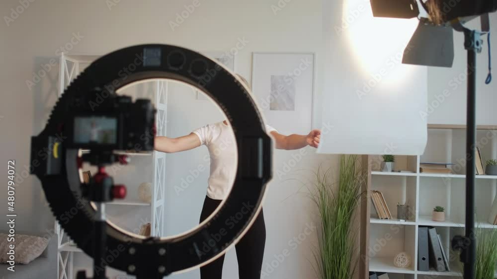 Fitness blog. Sportive woman. Video training. Smiling lady doing stretching workout for hands spinning around on photo camera in light room interior. Fitness blog. Sportive woman. Video training. Smiling lady doing stretching workout for hands spinning around on photo camera in light room interior.