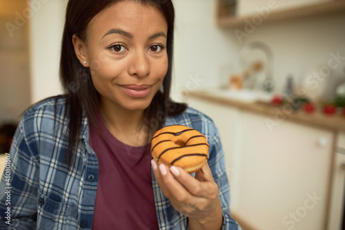 Happy beautiful charismatic African American woman holding tasty glazed doughnut  looking at camera with excitement  wearing blue plaid shirt  isolated on blurred background of kitchen counter-top