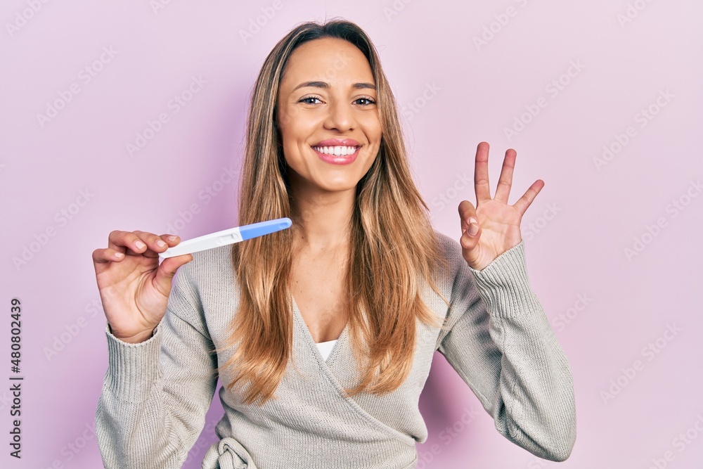 Beautiful hispanic woman holding pregnancy test result doing ok sign with fingers, smiling friendly gesturing excellent symbol Beautiful hispanic woman holding pregnancy test result doing ok sign with fingers, smiling friendly gesturing excellent symbol
