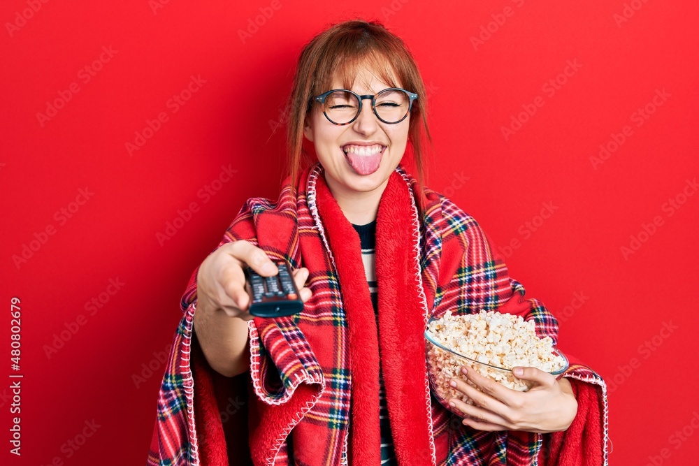 Redhead young woman eating popcorn using tv control sticking tongue out happy with funny expression. Redhead young woman eating popcorn using tv control sticking tongue out happy with funny expression.