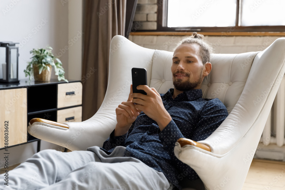 Φωτογραφία Happy handsome young hipster man relaxing in cozy armchair, involved in using mobile software applications, chatting in dating app or social network, playing games, spending time online or shopping