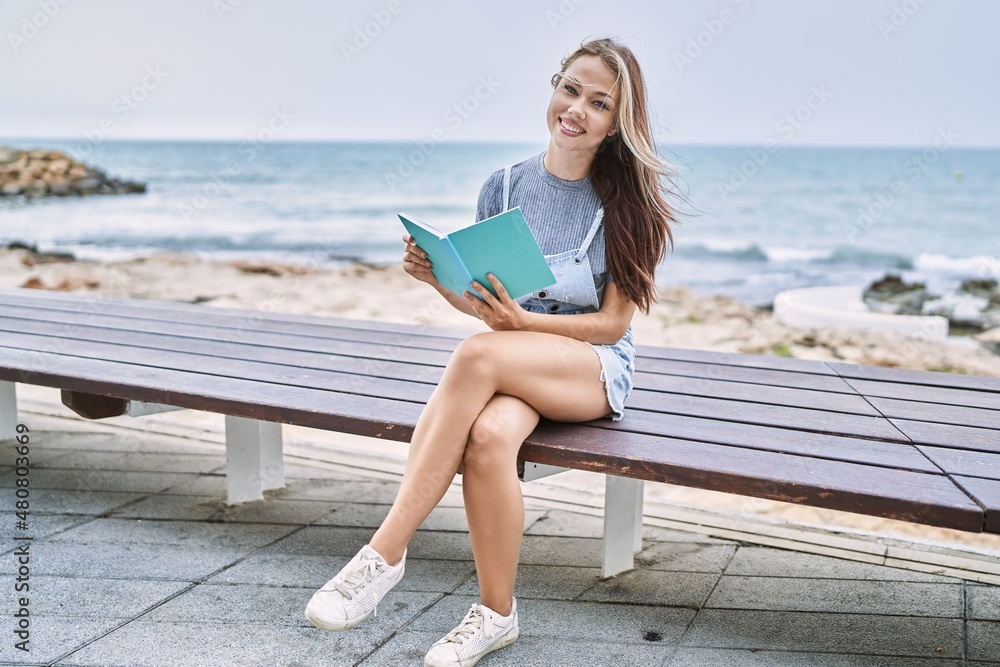 Young caucasian girl reading book sitting on the bench at the beach.