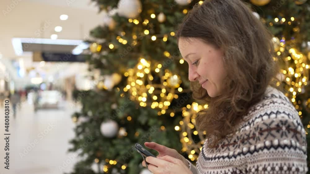 Young brunette curly haired woman in knitted Scandinavian sweater checks social media in phone walking around shopping mall past Christmas tree closeup Young brunette curly haired woman in knitted Scandinavian sweater checks social media in phone walking around shopping mall past Christmas tree closeup