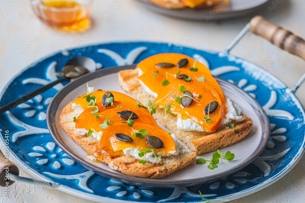Open sandwiches with curd cheese, persimmon slices and honey on a ceramic plate on a light concrete background. Italian food. Open sandwiches with curd cheese, persimmon slices and honey on a ceramic plate on a light concrete background. Italian food.