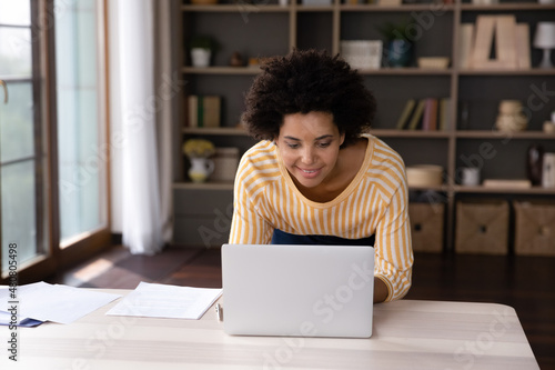 Happy concentrated millennial African American businesswoman looking at laptop screen, typng email communicating distantly, working on online project or preparing electronic document in modern office.