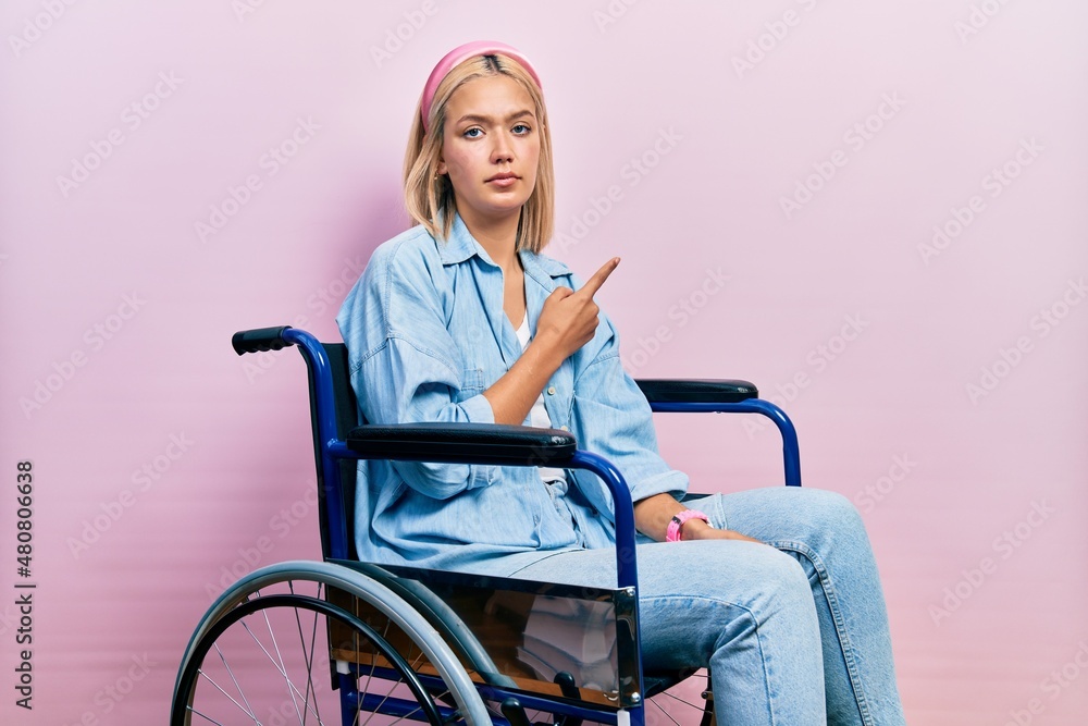 Beautiful blonde woman sitting on wheelchair pointing with hand finger to the side showing advertisement, serious and calm face Beautiful blonde woman sitting on wheelchair pointing with hand finger to the side showing advertisement, serious and calm face