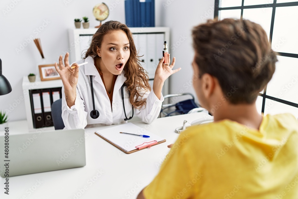 Fototapeta premium Young doctor woman showing electronic cigarette and normal cigarrete to patient afraid and shocked with surprise and amazed expression, fear and excited face.