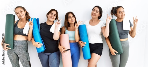 Group of women holding yoga mat standing over isolated background smiling looking to the camera showing fingers doing victory sign. number two.