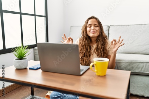 Beautiful hispanic woman using computer laptop at home relax and smiling with eyes closed doing meditation gesture with fingers. yoga concept.