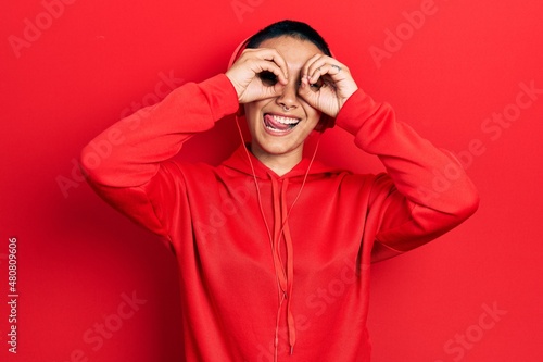 Beautiful hispanic woman with short hair listening to music using headphones doing ok gesture like binoculars sticking tongue out, eyes looking through fingers. crazy expression. © Krakenimages.com