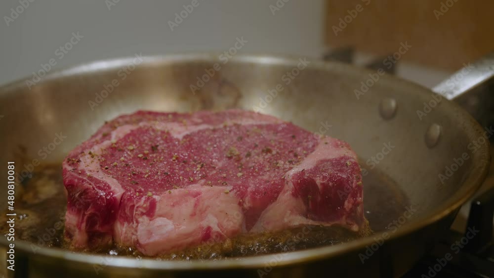 One juicy rib eye beef steak frying in high fat, in a pan. Red meat spiced with pepper, close up shot. One juicy rib eye beef steak frying in high fat, in a pan. Red meat spiced with pepper, close up shot.