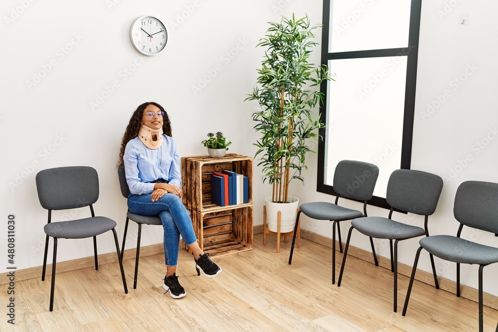 Fototapeta premium Young latin woman wearing neck collar sitting on chair at clinic waiting room