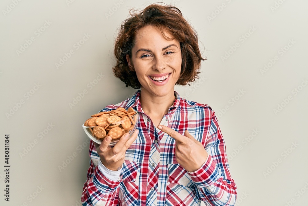 Young brunette woman holding bowl with salty biscuits smiling happy pointing with hand and finger Young brunette woman holding bowl with salty biscuits smiling happy pointing with hand and finger