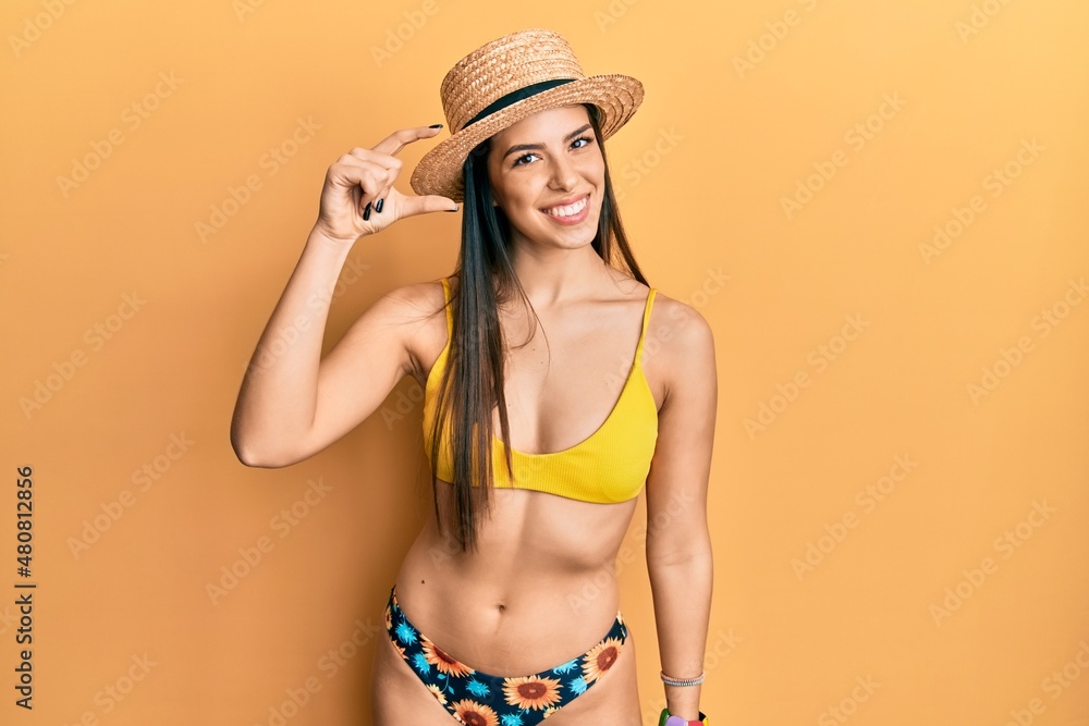 Young hispanic woman wearing bikini and summer hat smiling and confident gesturing with hand doing small size sign with fingers looking and the camera. measure concept. Young hispanic woman wearing bikini and summer hat smiling and confident gesturing with hand doing small size sign with fingers looking and the camera. measure concept.