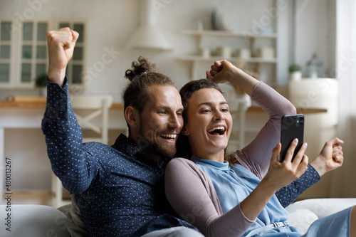 Obraz na plátně Laughing joyful bonding young family couple looking at smartphone screen, celebrating online lottery giveaway gambling game win, getting message with amazing news or big discount shopping promo code