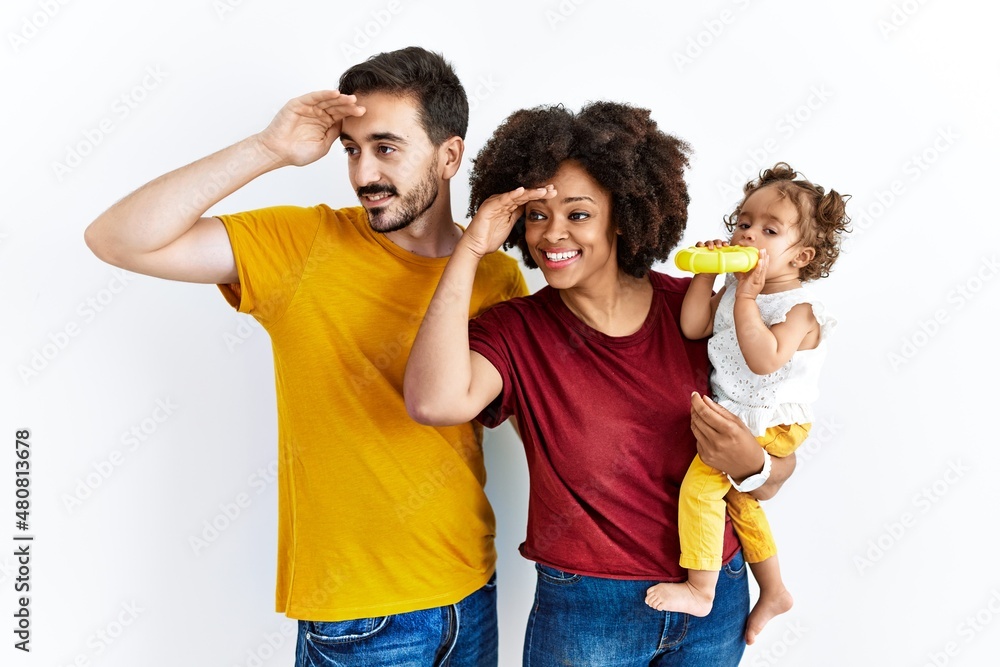 Interracial young family of black mother and hispanic father with daughter very happy and smiling looking far away with hand over head. searching concept. Interracial young family of black mother and hispanic father with daughter very happy and smiling looking far away with hand over head. searching concept.