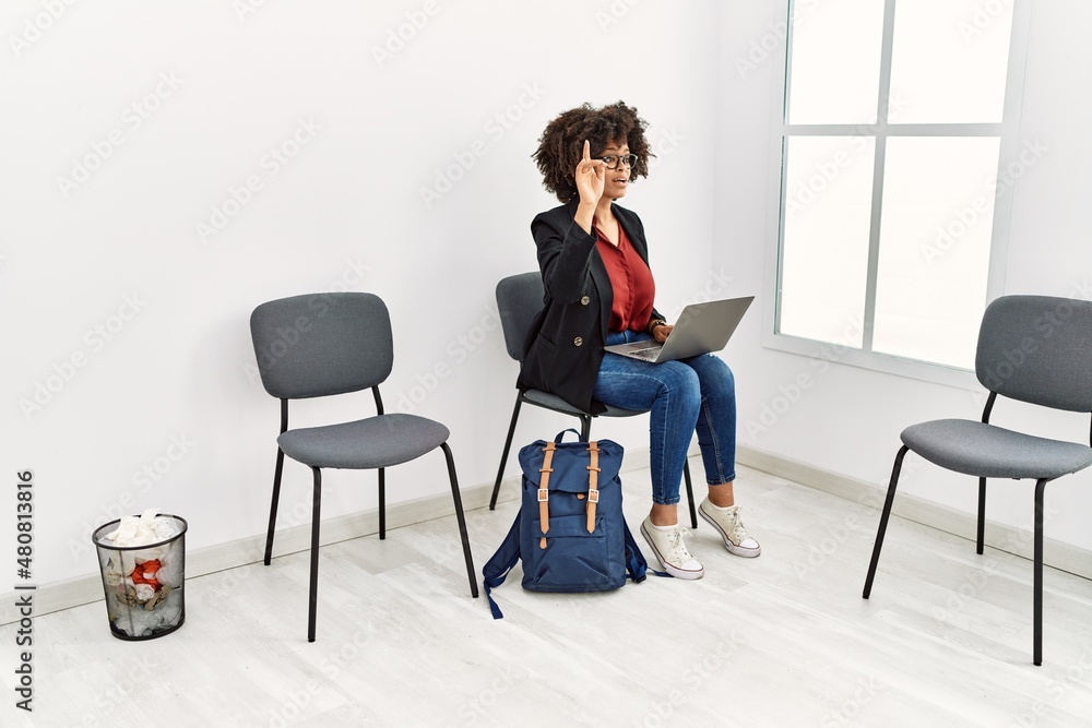 Fototapeta premium Young african american woman using laptop with finger raised up sitting on chair at waiting room