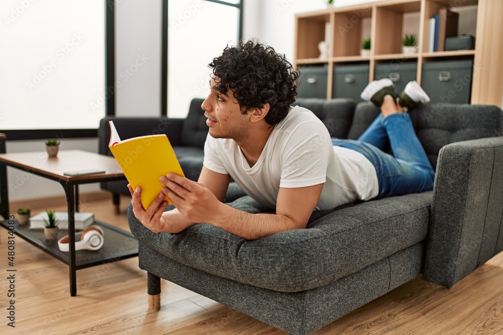 Young hispanic man reading book lying on the sofa at home.