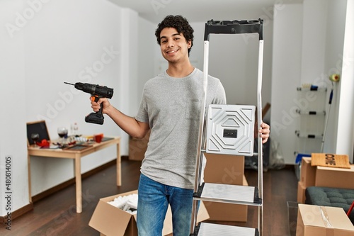 Young hispanic man smiling happy holding ladder and drill at new home. photo