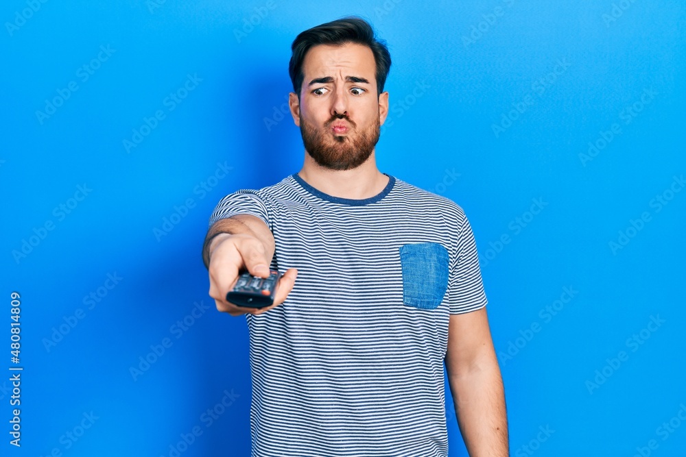 Handsome caucasian man with beard holding television remote control making fish face with mouth and squinting eyes, crazy and comical. Handsome caucasian man with beard holding television remote control making fish face with mouth and squinting eyes, crazy and comical.