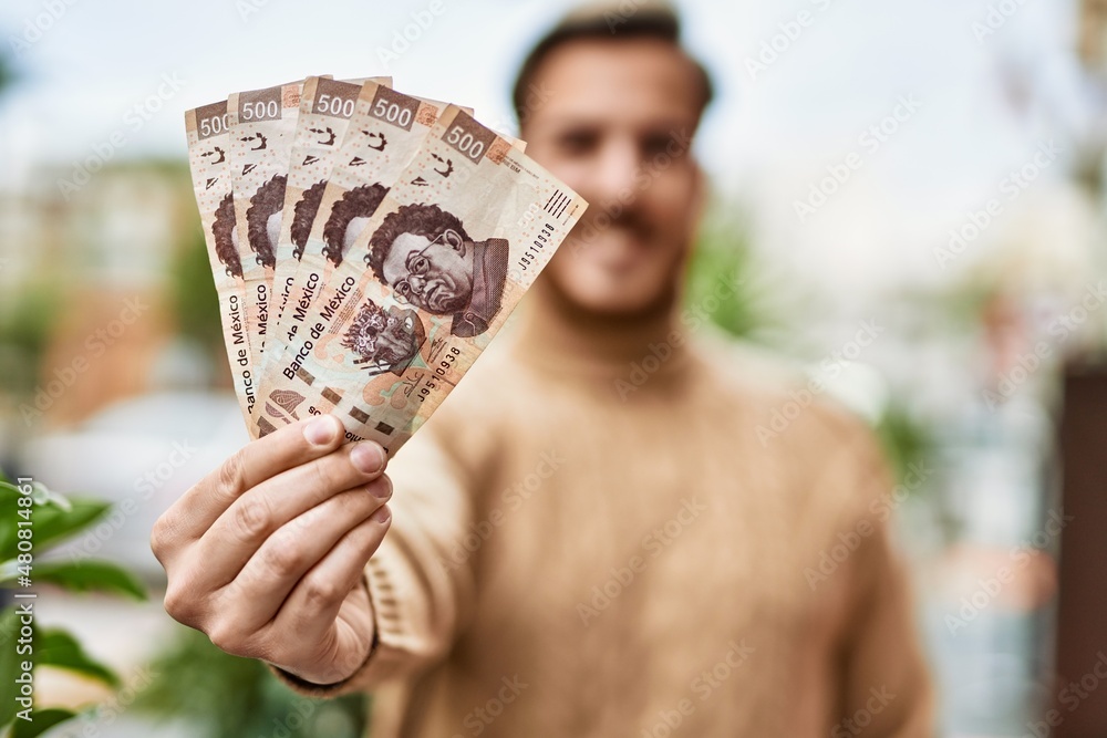 Young caucasian man smiling happy holding mexican pesos banknotes at the city. - obrazy, fototapety, plakaty 